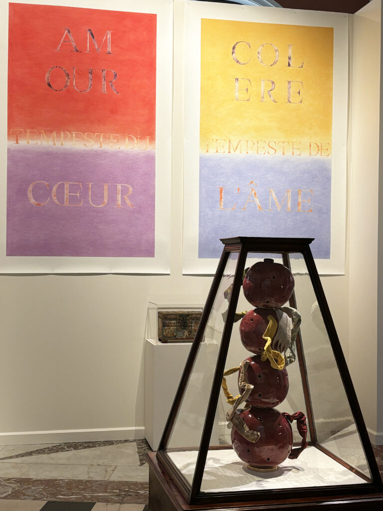 Two large typographic pastels (AM-OUR; COL-ÈRE, “Tempeste du cœur / de l’âme”) converse with the ceramic sculpture L’Énigme de Louise, bound with ribbons and shown in a pyramidal display case; in the background, an embroidered casket. Lise Terdjman’s exhibition Dearest Louise at the Musée de Picardie, Amiens (2025–2026).