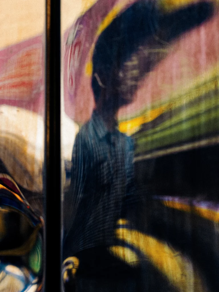 Reflet urbain déformé d’une silhouette dans une vitre, strates de couleurs chaudes et froides.
magazine art mag