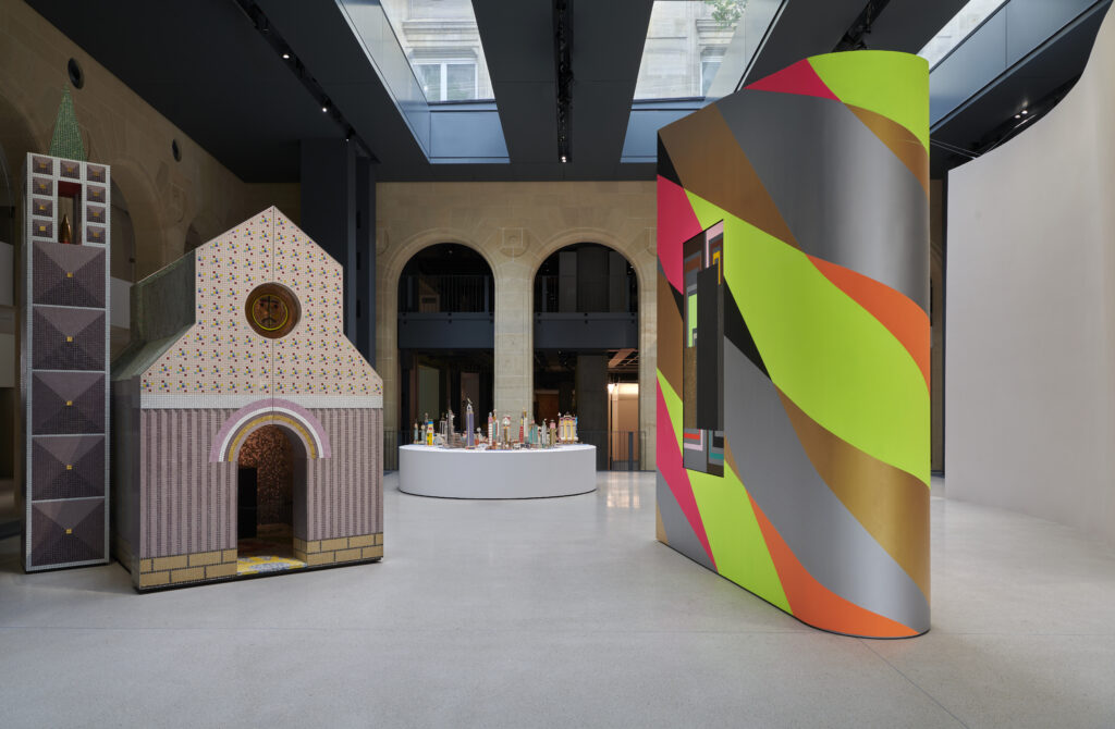 Exhibition view at the Fondation Cartier pour l’art contemporain, presenting works by Alessandro Mendini, Bodys Isek Kingelez and Peter Halley. On the left, a colourful architectural structure evokes a miniature church; in the centre, a circular table displays urban models; on the right, a large curved wall with fluorescent green, orange and grey geometric patterns. Natural light emphasises the architectural and dialogical dimension of the space. art mag magazine