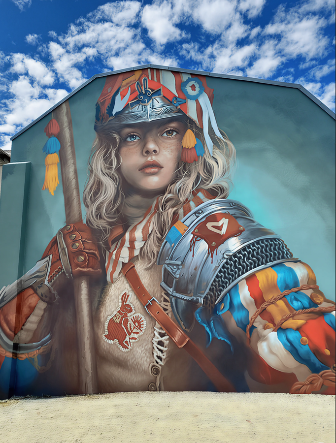 Fresque murale de l’artiste Arkane à Cransac-les-Thermes représentant une jeune fille en armure colorée, tenant une lance. Elle arbore des éléments médiévaux revisités avec des motifs contemporains, un blason en forme de cœur, des rubans tricolores, et un casque orné d’une fleur de lys. Le ciel bleu nuageux renforce la dimension héroïque et engagée de la scène.