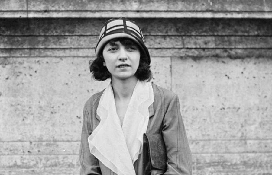 Portrait photographique d’Odette Pauvert, artiste peintre française, posant en manteau clair et chapeau à motifs géométriques devant un mur de pierre, années 1920.
