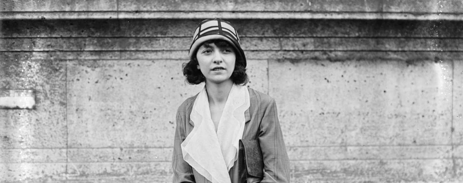 Portrait photographique d’Odette Pauvert, artiste peintre française, posant en manteau clair et chapeau à motifs géométriques devant un mur de pierre, années 1920.