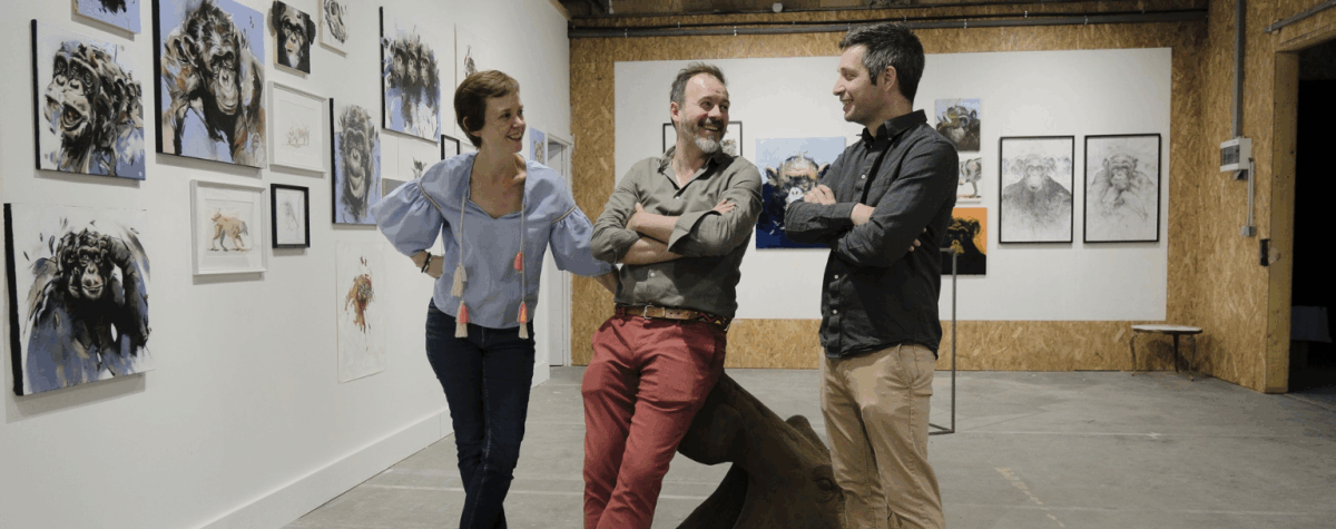 Sarah Scouarnec, Pierre-Jean Chabert et Thibault Jandot discutant devant une série d’œuvres animalières exposées aux Ateliers de la Morinerie à Saint-Pierre-des-Corps. À gauche, plusieurs peintures représentant des chimpanzés ; au centre, les trois artistes échangent dans une ambiance conviviale au cœur de l’espace d’exposition.