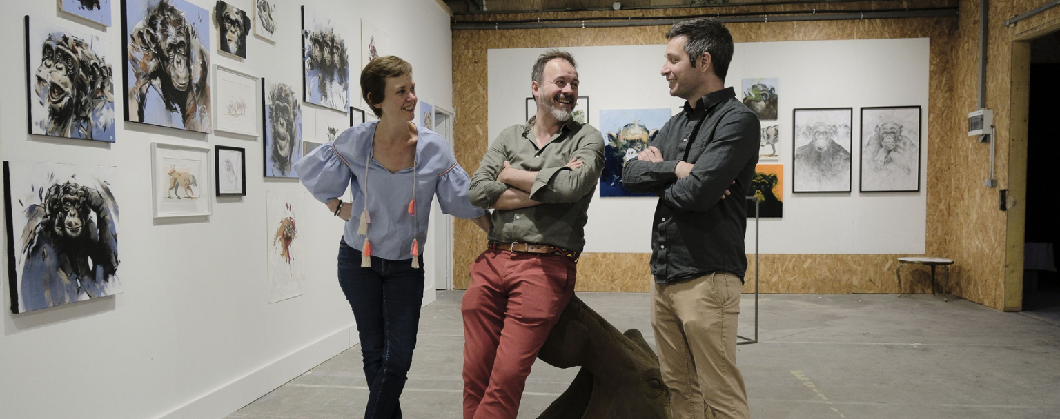 Sarah Scouarnec, Pierre-Jean Chabert et Thibault Jandot discutant devant une série d’œuvres animalières exposées aux Ateliers de la Morinerie à Saint-Pierre-des-Corps. À gauche, plusieurs peintures représentant des chimpanzés ; au centre, les trois artistes échangent dans une ambiance conviviale au cœur de l’espace d’exposition.