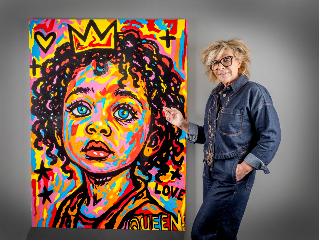 L’artiste Flo Muliardo pose à côté de l’une de ses toiles de la série Les Enfants Rois, représentant un jeune enfant aux grands yeux bleus, entouré de couleurs vives et contrastées — rouge, jaune, rose et bleu — surmonté d’une couronne dorée et des mots LOVE et QUEEN. La scène illustre l’univers expressif et coloré de l’artiste, célébrant la dignité et la force de l’enfance.