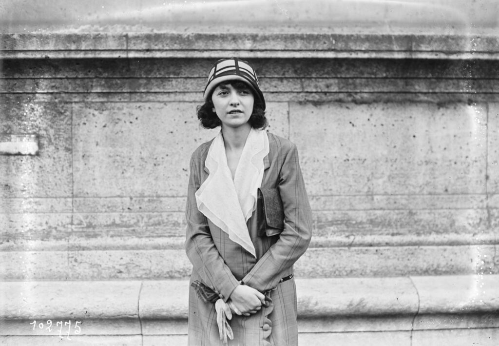 Portrait photographique d’Odette Pauvert, artiste peintre française, posant en manteau clair et chapeau à motifs géométriques devant un mur de pierre, années 1920.