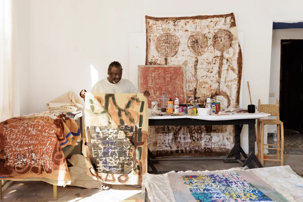 Sokey Edorh dans son atelier à Jardin Rouge, Marrakech (2025), préparant ses toiles pour l’exposition Waiting for Barbarians à la Fondation Montresso*. Magazine Art mag