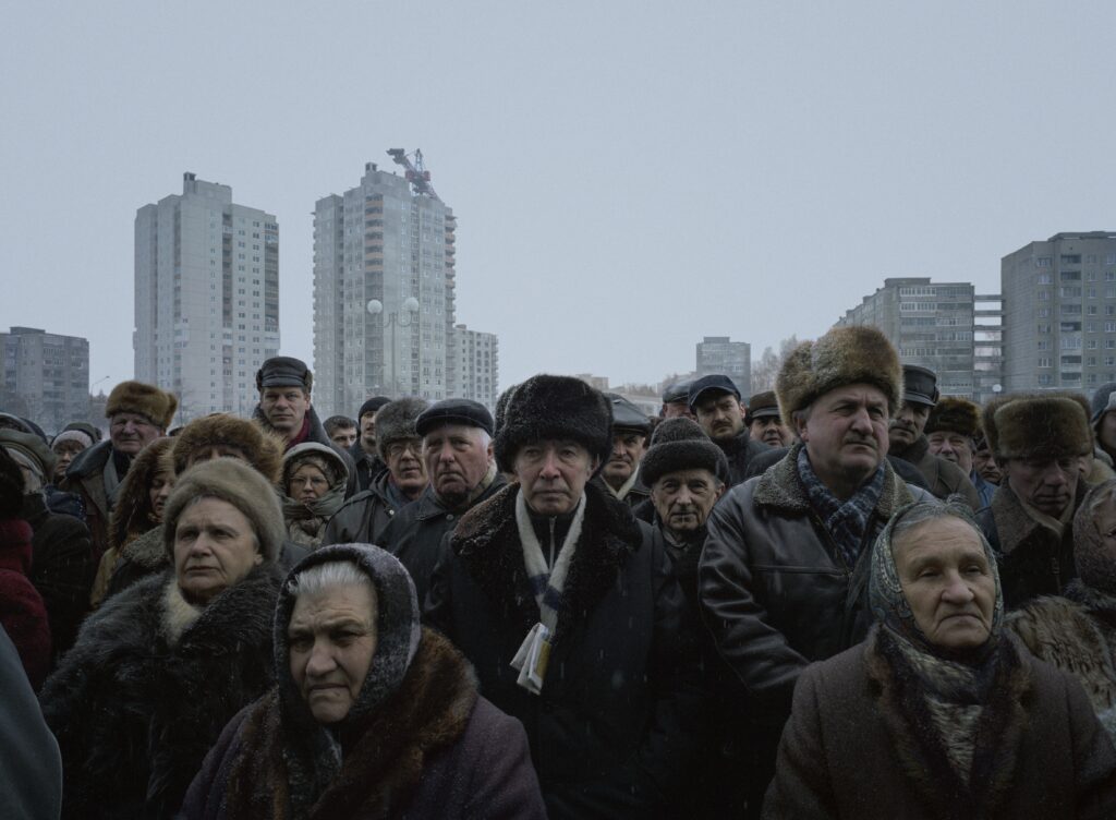 hotograph by Luc Delahaye depicting a crowd of elderly men and women gathered in the cold under a grey sky, in front of Eastern European apartment blocks. A solemn and silent scene about popular dignity and resistance. Exhibition The Noise of the World, Jeu de Paume, 2025. Magazine art mag