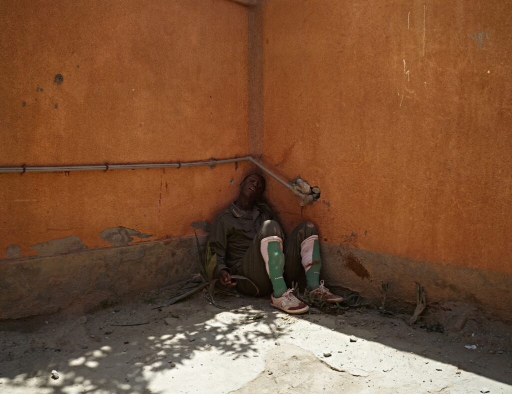 Photograph by Luc Delahaye showing a man lying against an orange wall, in a corner flooded with light. A striking work on human fragility and the harsh reality of war. Exhibition The Noise of the World, Jeu de Paume, 2025. Magazine Art Mag