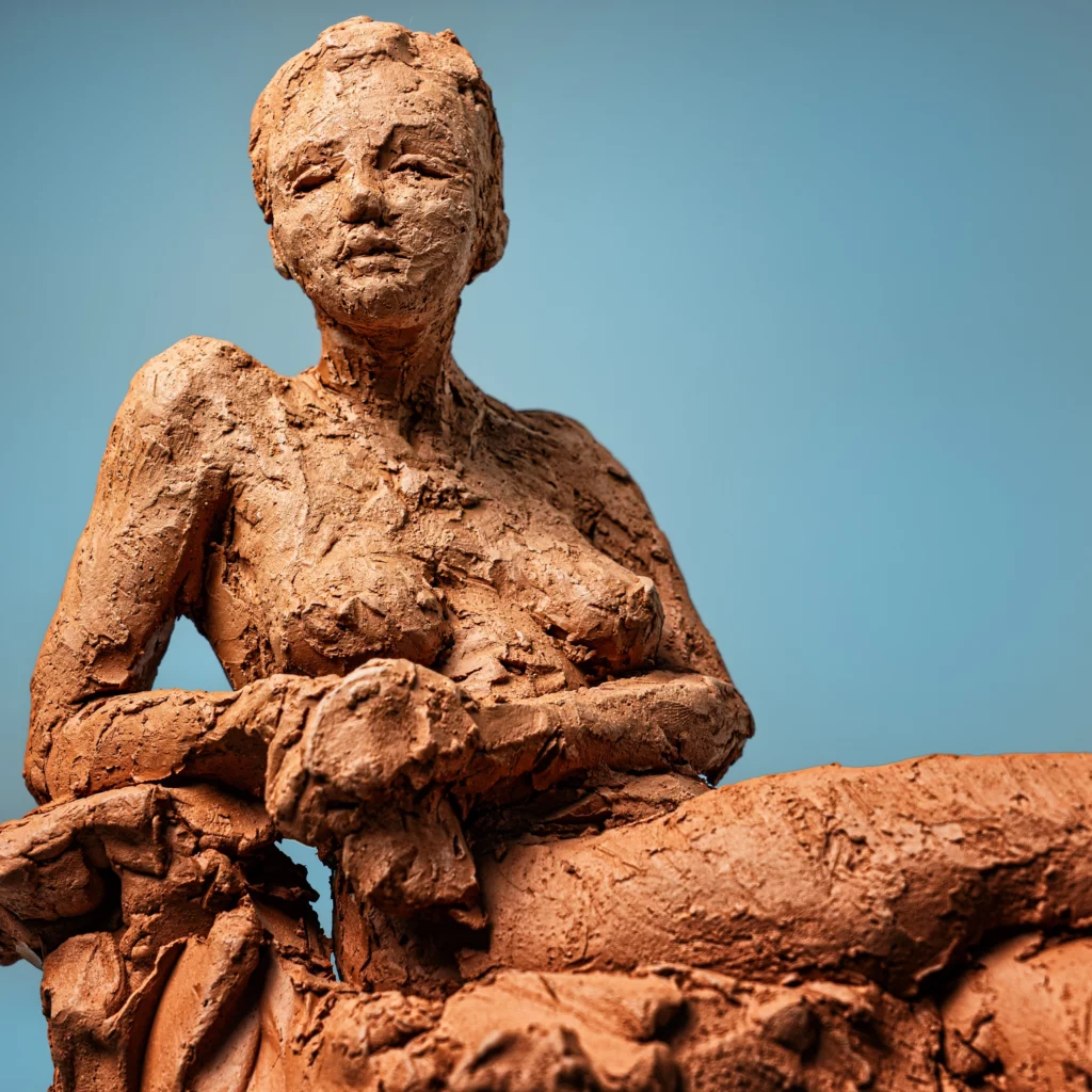 Sculpture d’un nu féminin en terre cuite, posture assise, textures rugueuses et expressives sur fond bleu.  Article Art mag