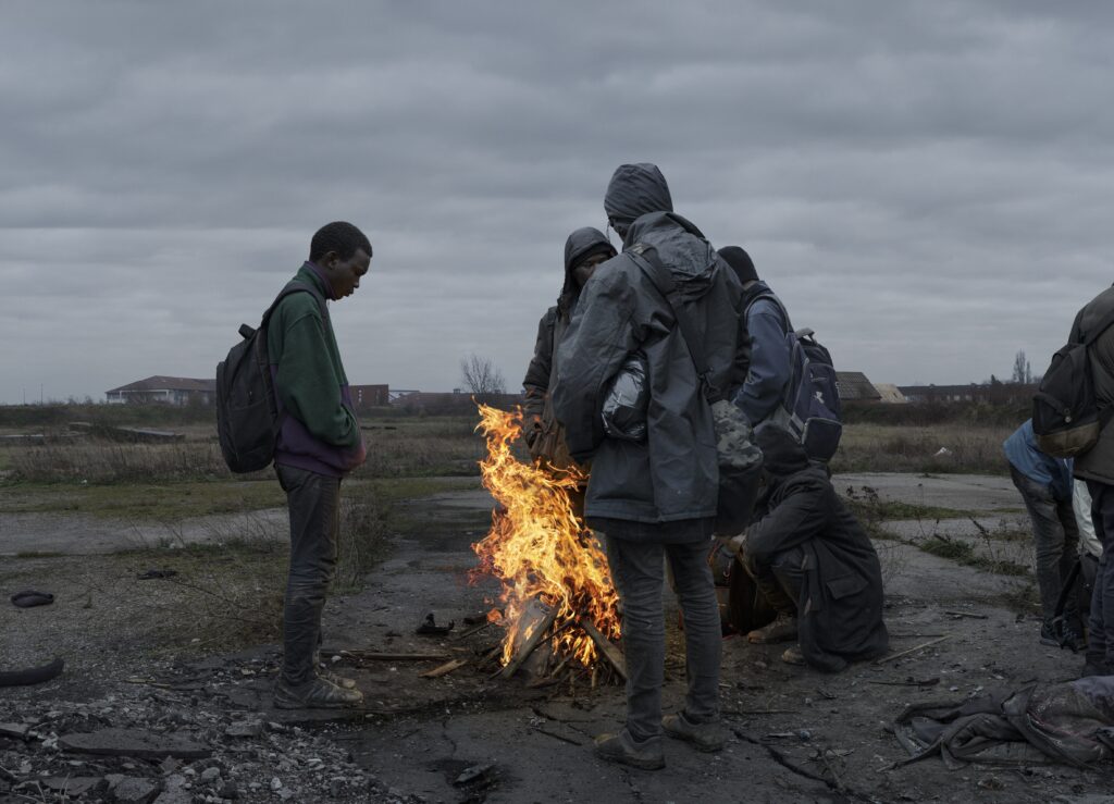 Photograph by Luc Delahaye showing a group of men gathered around a fire outdoors under a grey sky. A powerful image about survival, exile, and solidarity in a world in crisis. Exhibition The Noise of the World, Jeu de Paume, 2025. Article Art Mag