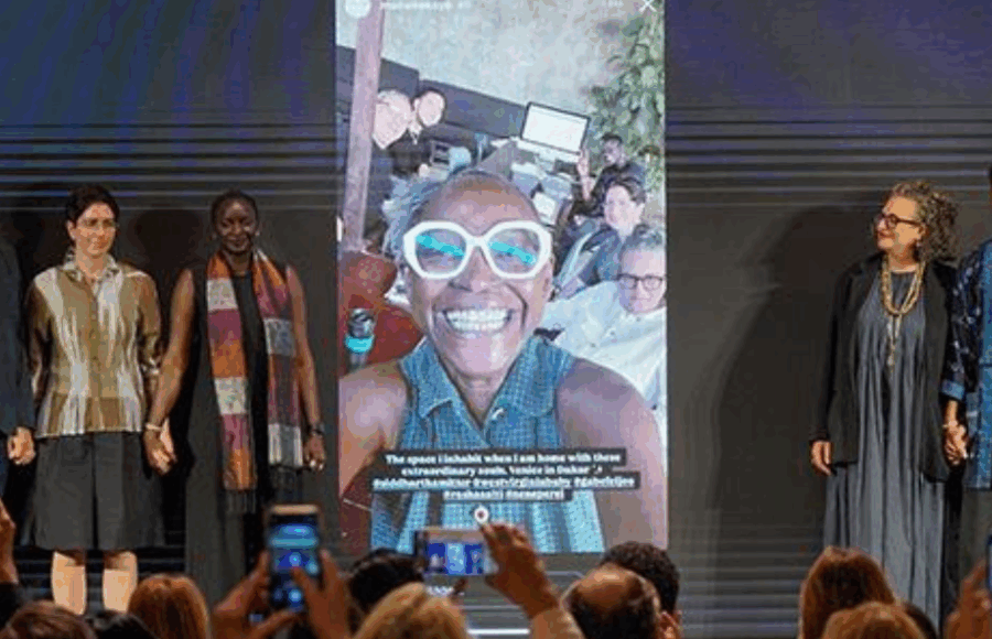 Six personnes sont alignées sur scène devant un public, se tenant par la main, avec en arrière-plan un grand écran montrant un portrait souriant de Koyo Kouoh entourée de son équipe. Cette image est présentée dans le cadre de la 61ᵉ Exposition internationale d’art, In Minor Keys, prévue du 9 mai au 22 novembre 2026, une édition conçue par la commissaire Koyo Kouoh. magazine art mag