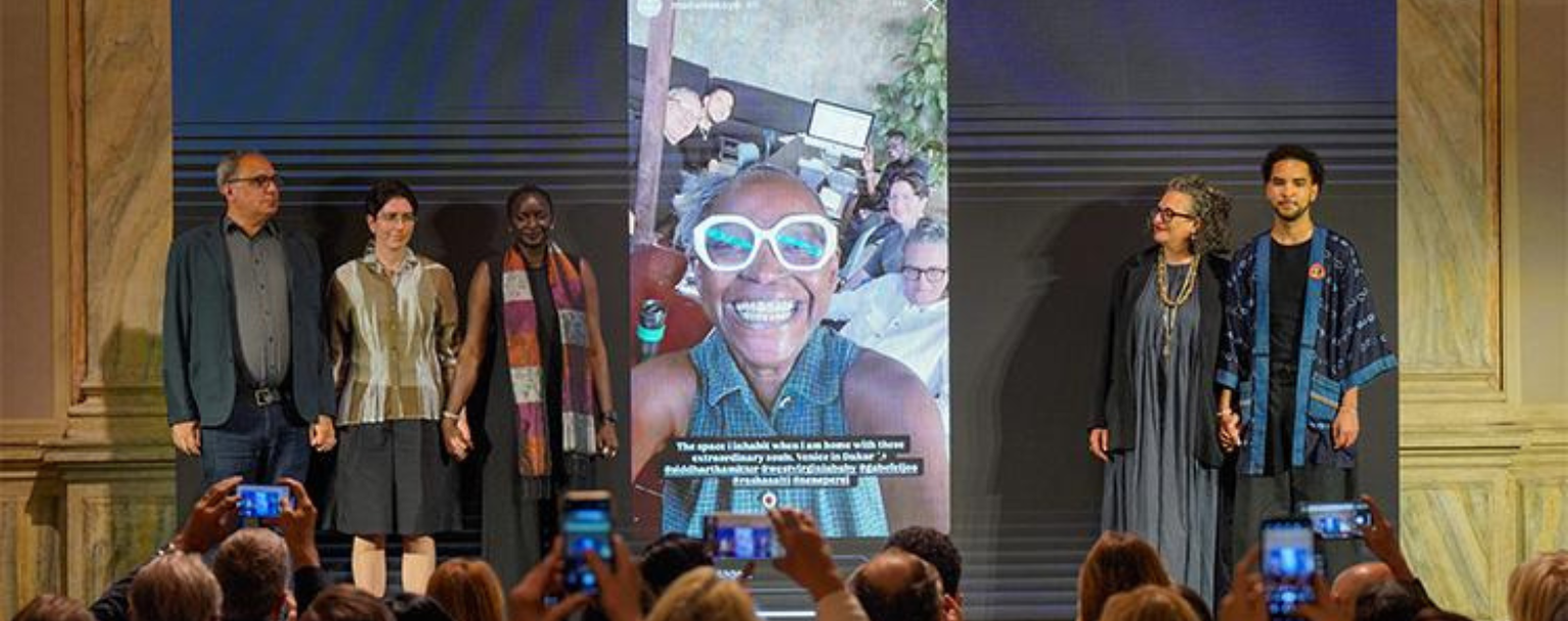 Six personnes sont alignées sur scène devant un public, se tenant par la main, avec en arrière-plan un grand écran montrant un portrait souriant de Koyo Kouoh entourée de son équipe. Cette image est présentée dans le cadre de la 61ᵉ Exposition internationale d’art, In Minor Keys, prévue du 9 mai au 22 novembre 2026, une édition conçue par la commissaire Koyo Kouoh. magazine art mag