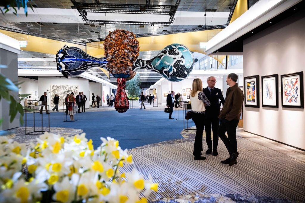 Vue générale de la BRAFA Art Fair montrant l’installation monumentale Valkyrie Leonie de Joana Vasconcelos suspendue au centre de l’allée principale. Des visiteurs échangent devant des œuvres contemporaines accrochées aux murs, dans un espace lumineux et élégant où fleurs, stands et galeries se déploient. Photographie d’ambiance illustrant la scénographie et l’atmosphère de la foire d’art de Bruxelles.