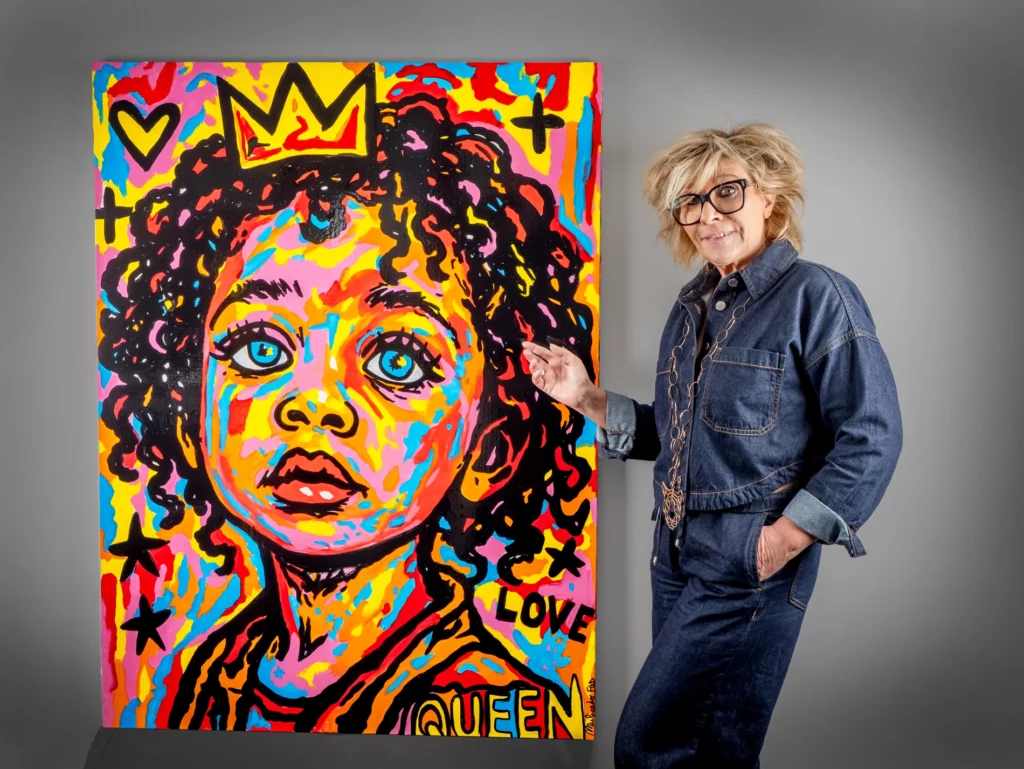 L’artiste Flo Muliardo pose à côté de l’une de ses toiles de la série Les Enfants Rois, représentant un jeune enfant aux grands yeux bleus, entouré de couleurs vives et contrastées — rouge, jaune, rose et bleu — surmonté d’une couronne dorée et des mots LOVE et QUEEN. La scène illustre l’univers expressif et coloré de l’artiste, célébrant la dignité et la force de l’enfance. Art Mag