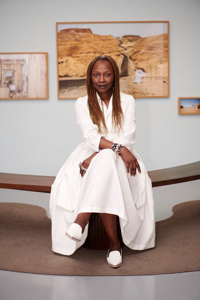 Commissaire de l’Exposition internationale d’art de la Biennale de Venise 2026 assise sur un banc en bois dans une galerie, vêtue d’une longue robe blanche et chaussée de mocassins blancs. Elle pose calmement, les mains croisées, avec de longues tresses. En arrière-plan, plusieurs photographies de paysages désertiques sont accrochées au mur magazine art mag