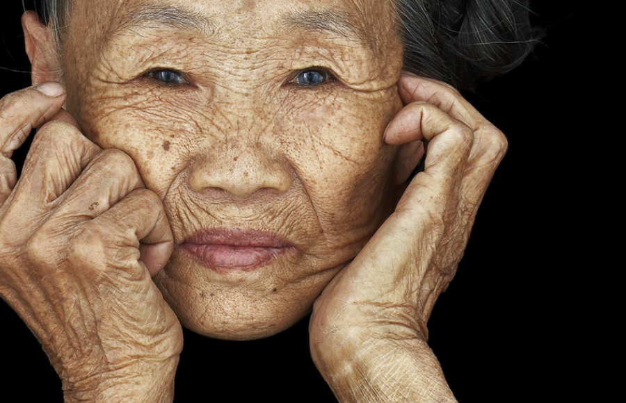 Portrait en gros plan d’une femme âgée sur fond noir, mains posées sur le visage – photographie d’Antoine Schneck, série « Chen Nai Ben », présentée à Photo Days 2025, Galerie Harcourt, partenaire de Paris Photo – magazine ART MAG.