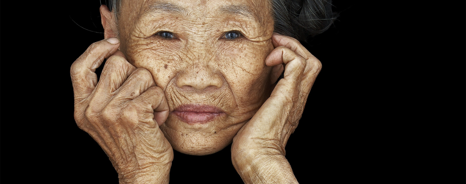 Portrait en gros plan d’une femme âgée sur fond noir, mains posées sur le visage – photographie d’Antoine Schneck, série « Chen Nai Ben », présentée à Photo Days 2025, Galerie Harcourt, partenaire de Paris Photo – magazine ART MAG.