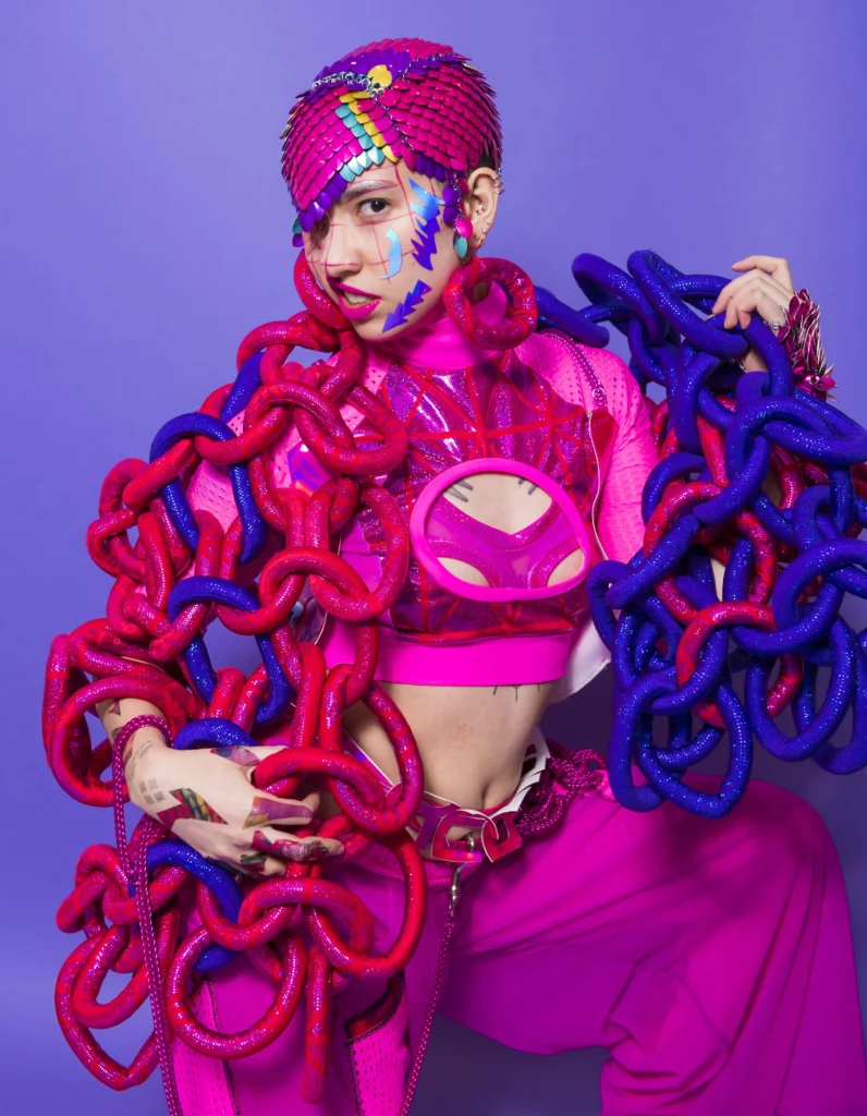 Modèle portant une création colorée et inclusive de Rebirth Garments, photographiée sur fond violet, célébrant la diversité des corps et la mode queer dans l’exposition Design and Disability au V&A Museum. publié magazine art mag