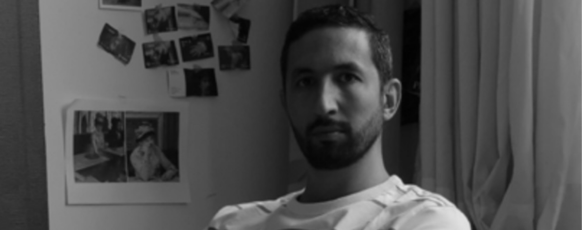 Black and white portrait of Bilal Hamdad in his studio, sitting with his arms crossed in front of a wall covered with photographs, illustrating the creative process of the contemporary artist exhibited at the Petit Palais in Paris.