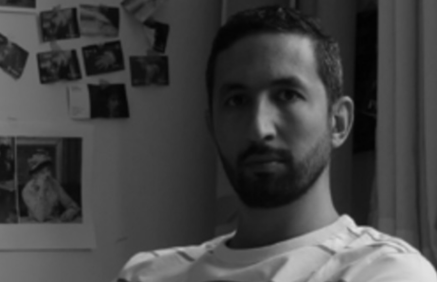 Black and white portrait of Bilal Hamdad in his studio, sitting with his arms crossed in front of a wall covered with photographs, illustrating the creative process of the contemporary artist exhibited at the Petit Palais in Paris.