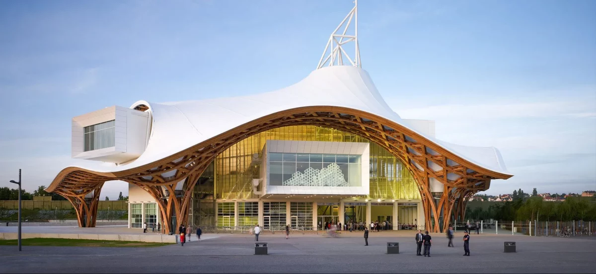Photograph of a contemporary building with organic architecture, featuring a large undulating white roof supported by a curved wooden structure, with visitors visible at the entrance.