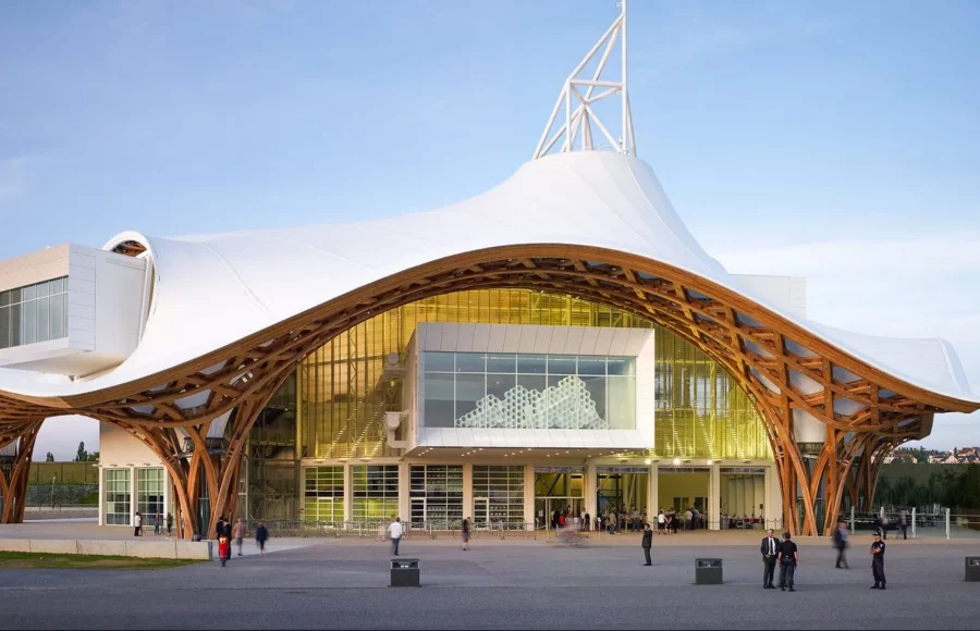 Photograph of a contemporary building with organic architecture, featuring a large undulating white roof supported by a curved wooden structure, with visitors visible at the entrance.