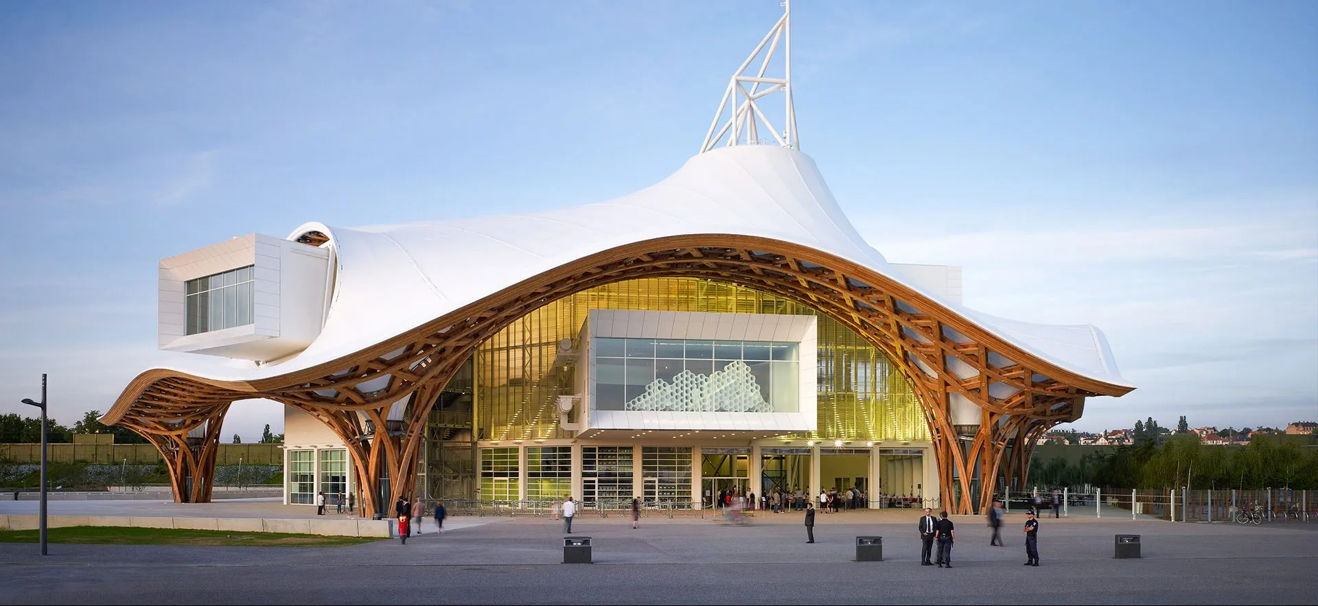 Photograph of a contemporary building with organic architecture, featuring a large undulating white roof supported by a curved wooden structure, with visitors visible at the entrance.