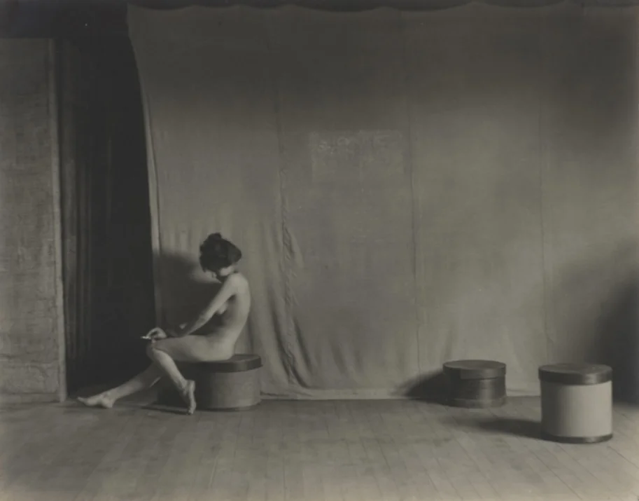 Photographie en noir et blanc d’une personne nue assise sur un tabouret, tournée vers la gauche dans un vaste studio. La figure, légèrement courbée et tenant un objet fin dans la main, est placée près d’un rideau sombre qui occupe presque toute la largeur de l’image. Le sol en bois et deux autres tabourets espacés complètent la scène minimaliste.