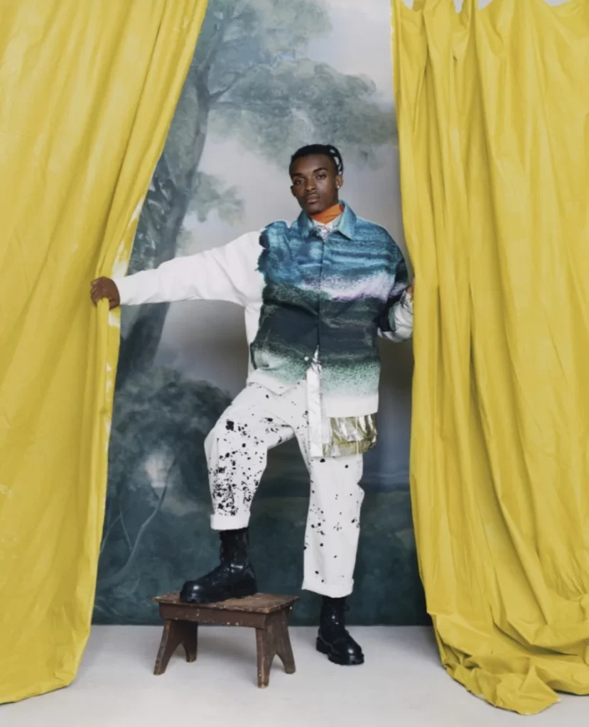 Colour photograph showing a person standing between two large yellow curtains pulled apart. She is posing with one foot on a small wooden stool, wearing a blue and green gradient jacket, white trousers with black spots, and black boots. In the background, a painted backdrop depicts a wooded landscape.