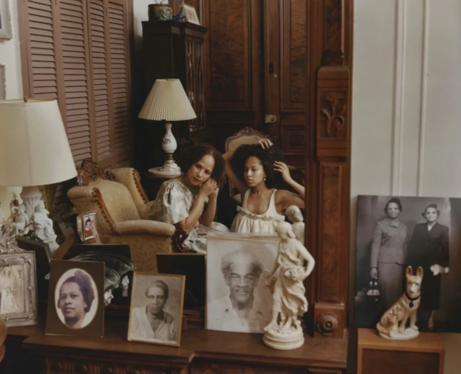 Indoor photograph showing two women seated in front of a large mirror decorated with carved wood. One, wearing a light-coloured dress, sits upright and adjusts her hair, while the other, seated beside her, observes her reflection. Around the mirror, numerous framed antique portraits are arranged on a piece of furniture, along with decorative statuettes and a lamp. The atmosphere evokes an intimate space steeped in family history.