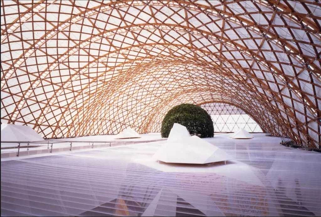 Structure architecturale conçue par Shigeru Ban, composée d’une vaste charpente en bois tressé formant une couverture légère et lumineuse, vue intérieure.