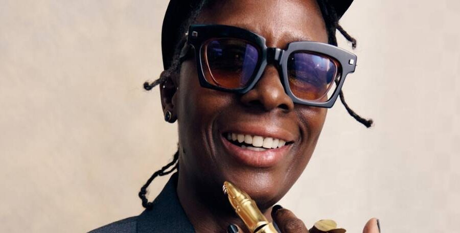 Portrait d'une personne souriante vêtue d'une veste de costume sombre, d'une casquette noire et de grandes lunettes rectangulaires. Elle porte des bijoux en or, notamment une bague articulée et un bracelet, et pose avec une main près du visage sur un fond neutre.