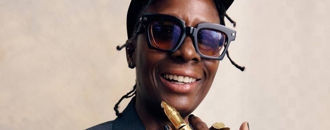 Portrait d'une personne souriante vêtue d'une veste de costume sombre, d'une casquette noire et de grandes lunettes rectangulaires. Elle porte des bijoux en or, notamment une bague articulée et un bracelet, et pose avec une main près du visage sur un fond neutre.