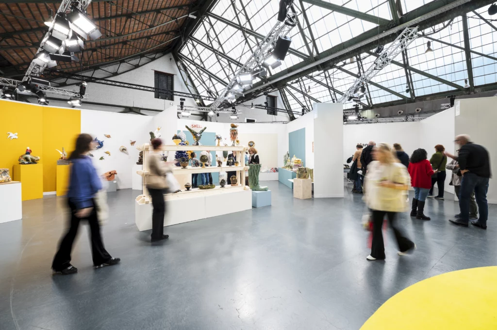 Exposition de céramique contemporaine à ceramic brussels ; visiteurs observant des sculptures et installations en argile sur socles, œuvres murales en céramique dans un espace d’art à Tour & Taxis.