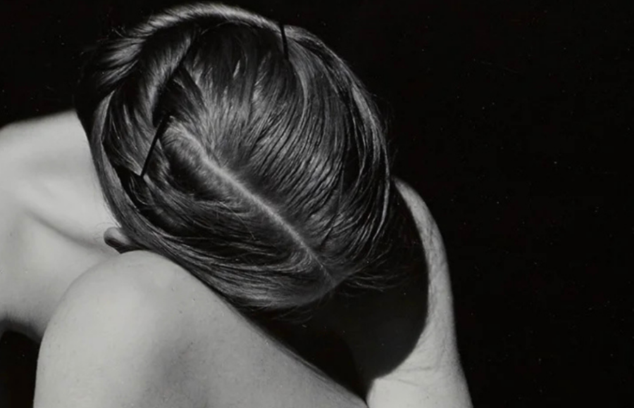 Photographie en noir et blanc représentant une personne nue assise au sol, le corps replié et la tête inclinée vers les genoux. Les bras entourent les jambes, formant une composition sculpturale où la lumière met en valeur les lignes, ombres et textures du corps.