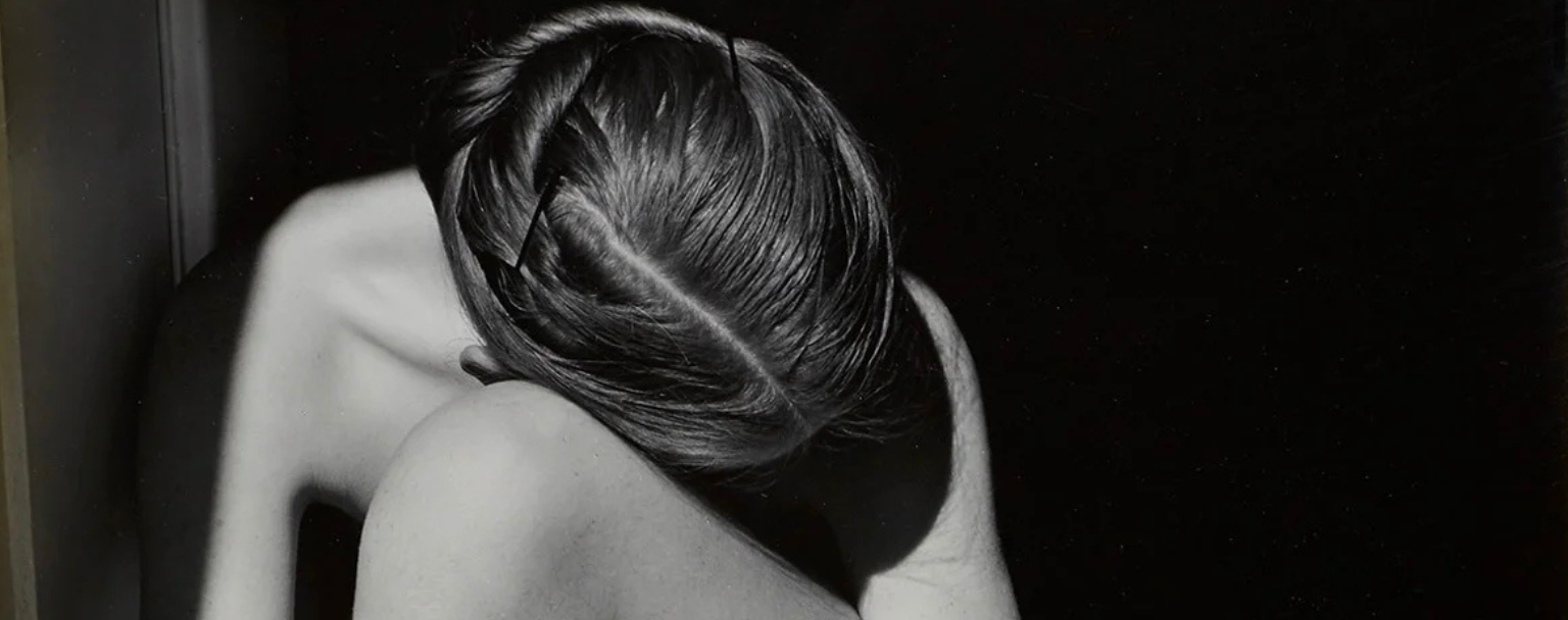 Photographie en noir et blanc représentant une personne nue assise au sol, le corps replié et la tête inclinée vers les genoux. Les bras entourent les jambes, formant une composition sculpturale où la lumière met en valeur les lignes, ombres et textures du corps.