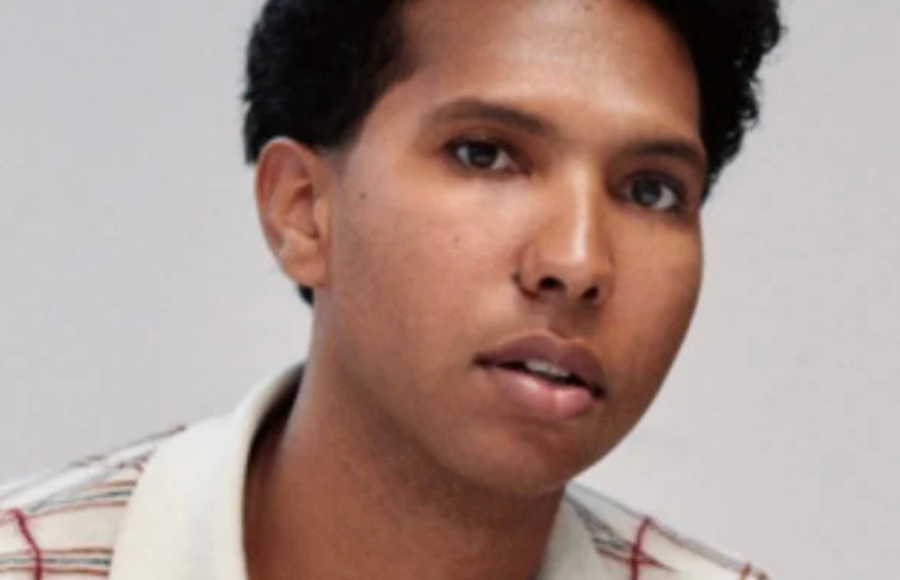 Colour portrait of a person sitting cross-legged on a white surface. They are wearing a cream and red checked top and blue jeans. Their hands are resting in front of them, with a bracelet and ring visible. The person is looking slightly to the right, with a calm expression.