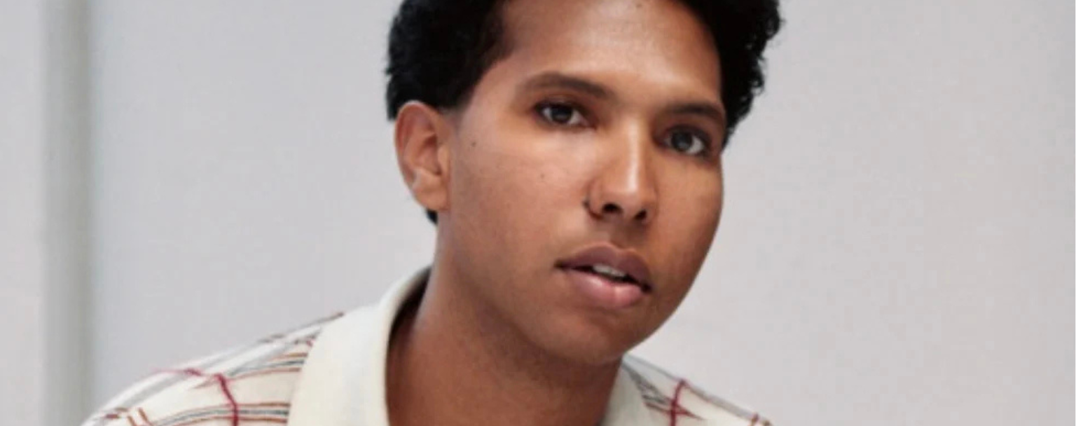 Colour portrait of a person sitting cross-legged on a white surface. They are wearing a cream and red checked top and blue jeans. Their hands are resting in front of them, with a bracelet and ring visible. The person is looking slightly to the right, with a calm expression.