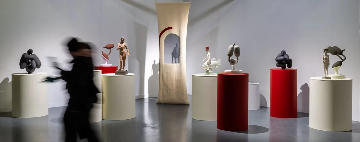 Vue d’exposition de ceramic brussels 2025 présentant des sculptures céramiques contemporaines sur socles blancs et rouges dans un espace lumineux, avec un visiteur en mouvement au premier plan.
