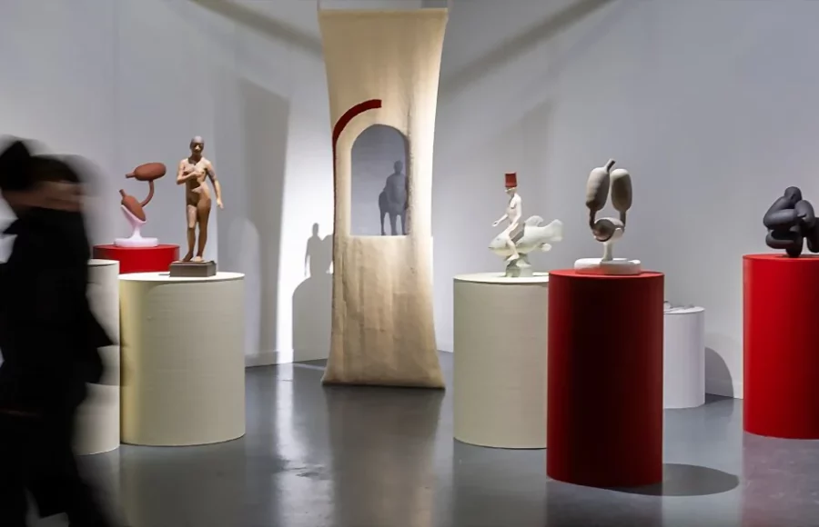 Vue d’exposition de ceramic brussels 2025 présentant des sculptures céramiques contemporaines sur socles blancs et rouges dans un espace lumineux, avec un visiteur en mouvement au premier plan.