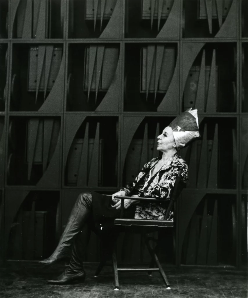 Figure seated in front of a monumental wooden wall sculpture, composed of geometric modules and vertical elements, photographed in black and white, illustrating the immersive and architectural sculptural universe associated with the exhibition ‘Mrs. N's Palace’ and the aesthetics of 20th-century environmental sculpture.