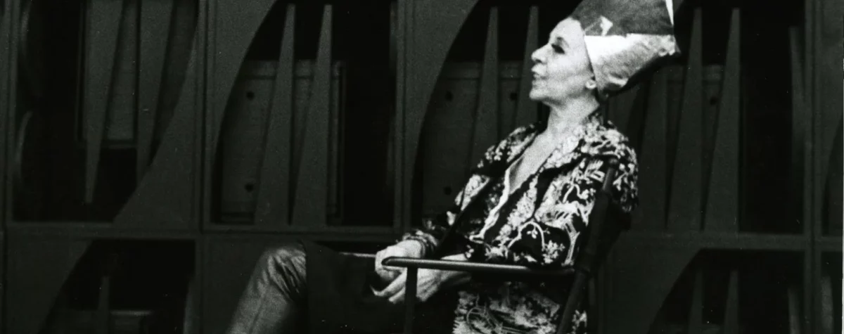 Figure seated in front of a monumental wooden wall sculpture, composed of geometric modules and vertical elements, photographed in black and white, illustrating the immersive and architectural sculptural universe associated with the exhibition ‘Mrs. N's Palace’ and the aesthetics of 20th-century environmental sculpture.