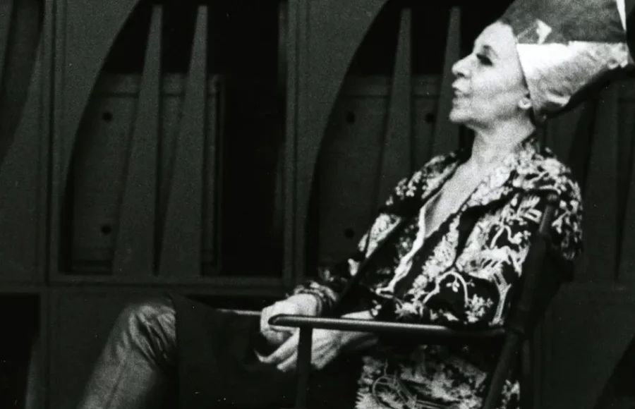 Figure seated in front of a monumental wooden wall sculpture, composed of geometric modules and vertical elements, photographed in black and white, illustrating the immersive and architectural sculptural universe associated with the exhibition ‘Mrs. N's Palace’ and the aesthetics of 20th-century environmental sculpture.