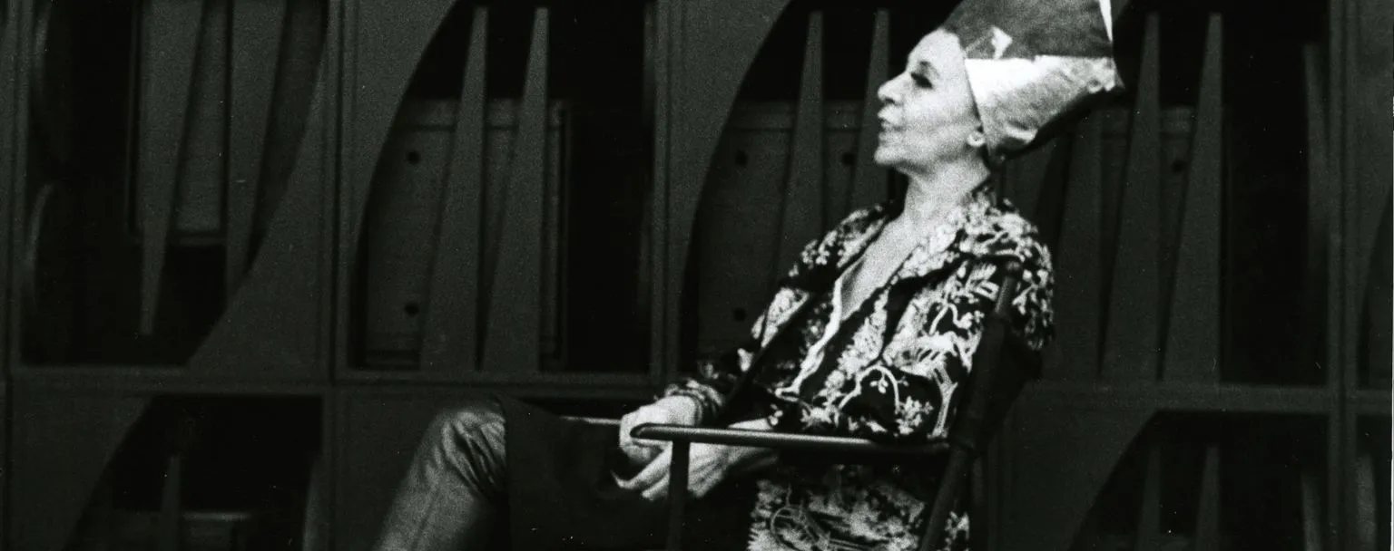 Figure seated in front of a monumental wooden wall sculpture, composed of geometric modules and vertical elements, photographed in black and white, illustrating the immersive and architectural sculptural universe associated with the exhibition ‘Mrs. N's Palace’ and the aesthetics of 20th-century environmental sculpture.