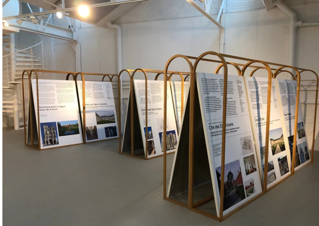 Vue intérieure d’une salle d’exposition lumineuse, avec plusieurs structures en bois en forme d’arceaux présentant des panneaux explicatifs inclinés. Les panneaux comportent des textes et des photographies sur le patrimoine architectural des Hauts-de-France. L’espace est épuré, avec murs blancs, sol gris et éclairage zénithal.