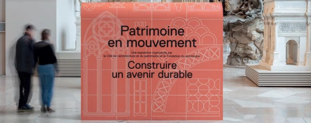 Texte alternatif proposé : « Panneau d’exposition rouge au centre d’un musée, portant le texte “Patrimoine en mouvement – Construire un avenir durable”, avec des motifs architecturaux dessinés en lignes claires. Deux visiteurs flous passent sur la gauche, et des éléments sculpturaux et architecturaux sont visibles à l’arrière-plan.