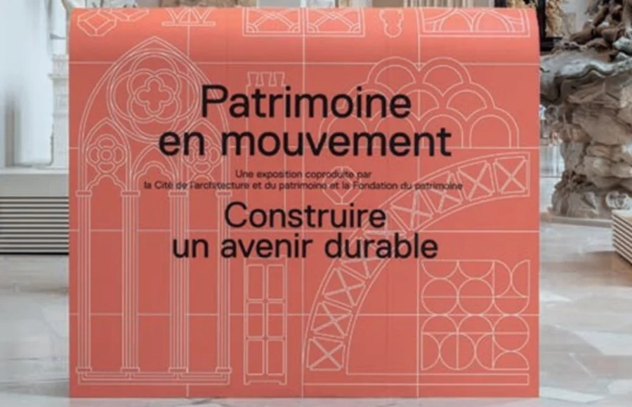 Texte alternatif proposé : « Panneau d’exposition rouge au centre d’un musée, portant le texte “Patrimoine en mouvement – Construire un avenir durable”, avec des motifs architecturaux dessinés en lignes claires. Deux visiteurs flous passent sur la gauche, et des éléments sculpturaux et architecturaux sont visibles à l’arrière-plan.
