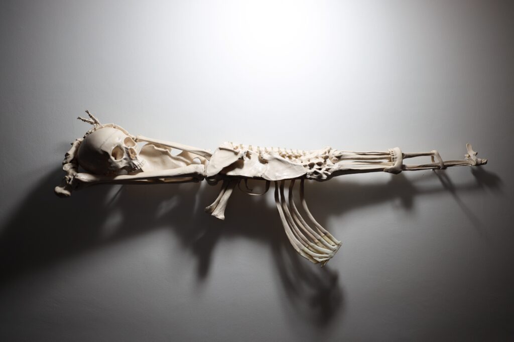 Anatomy of Authority – Julien Magic, sculpture contemporaine représentant un squelette humain fusionné avec une Kalachnikov, symbole du pouvoir et de la violence.