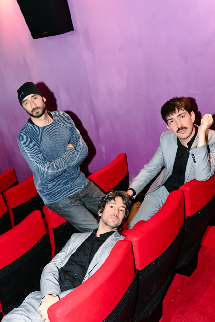Portrait de trois artistes de No Show, installés parmi les fauteuils rouges d’une salle de spectacle aux murs violets. Photographie de Sepideh Farvardin.